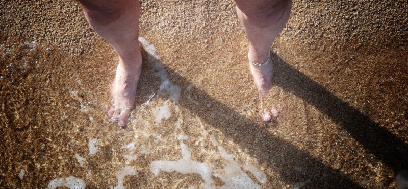 Deux pieds dans le sable, caressés par la mer. On distingue un bracelet qui ceint la cheville gauche.